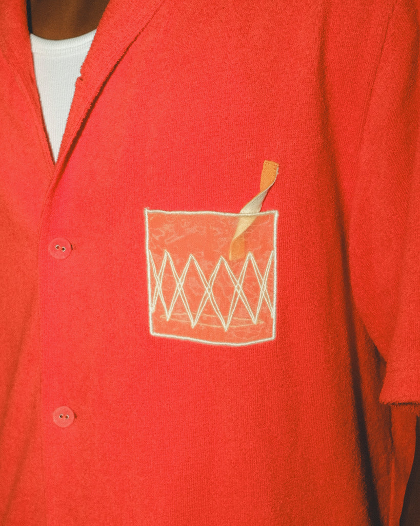 Close-up of a red cabana shirt showing the embroidered Negroni glass on the chest pocket, featuring a transparent mesh panel and stitched orange garnish detail.