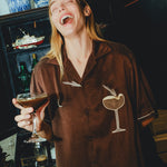 Woman wearing brown espresso martini shirt, while holding a drink in her hand and laughing.