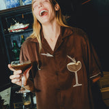 Woman wearing brown espresso martini shirt, while holding a drink in her hand and laughing.