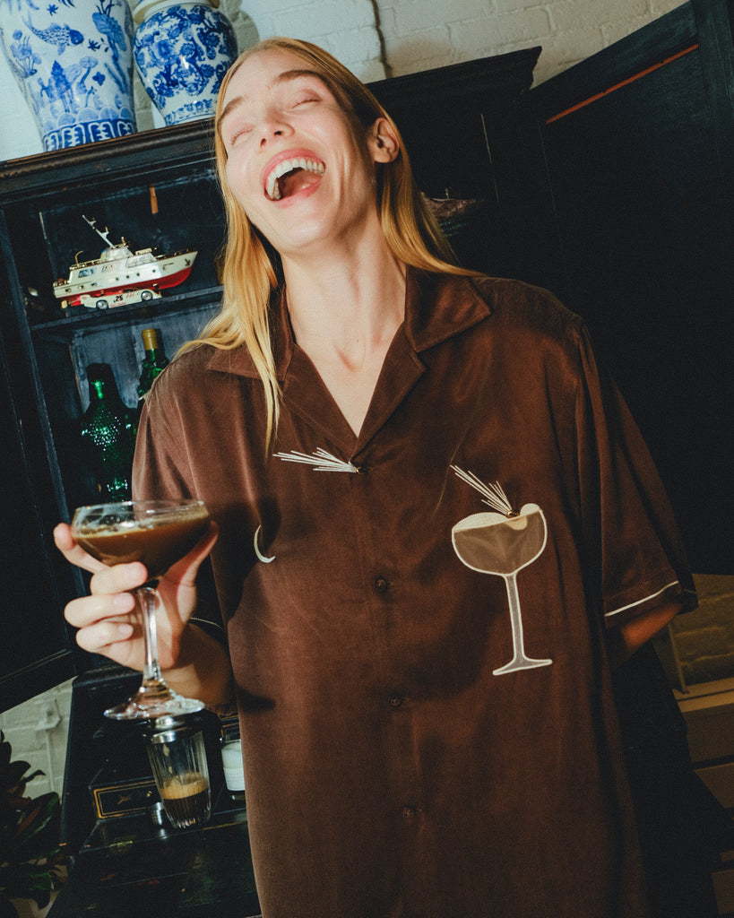 Woman wearing brown espresso martini shirt, while holding a drink in her hand and laughing.