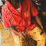 Model wearing a red cabana shirt with twisted appliqué stripes and an embroidered Negroni glass on the chest pocket, seated with confetti scattered across their clothing and surroundings. 