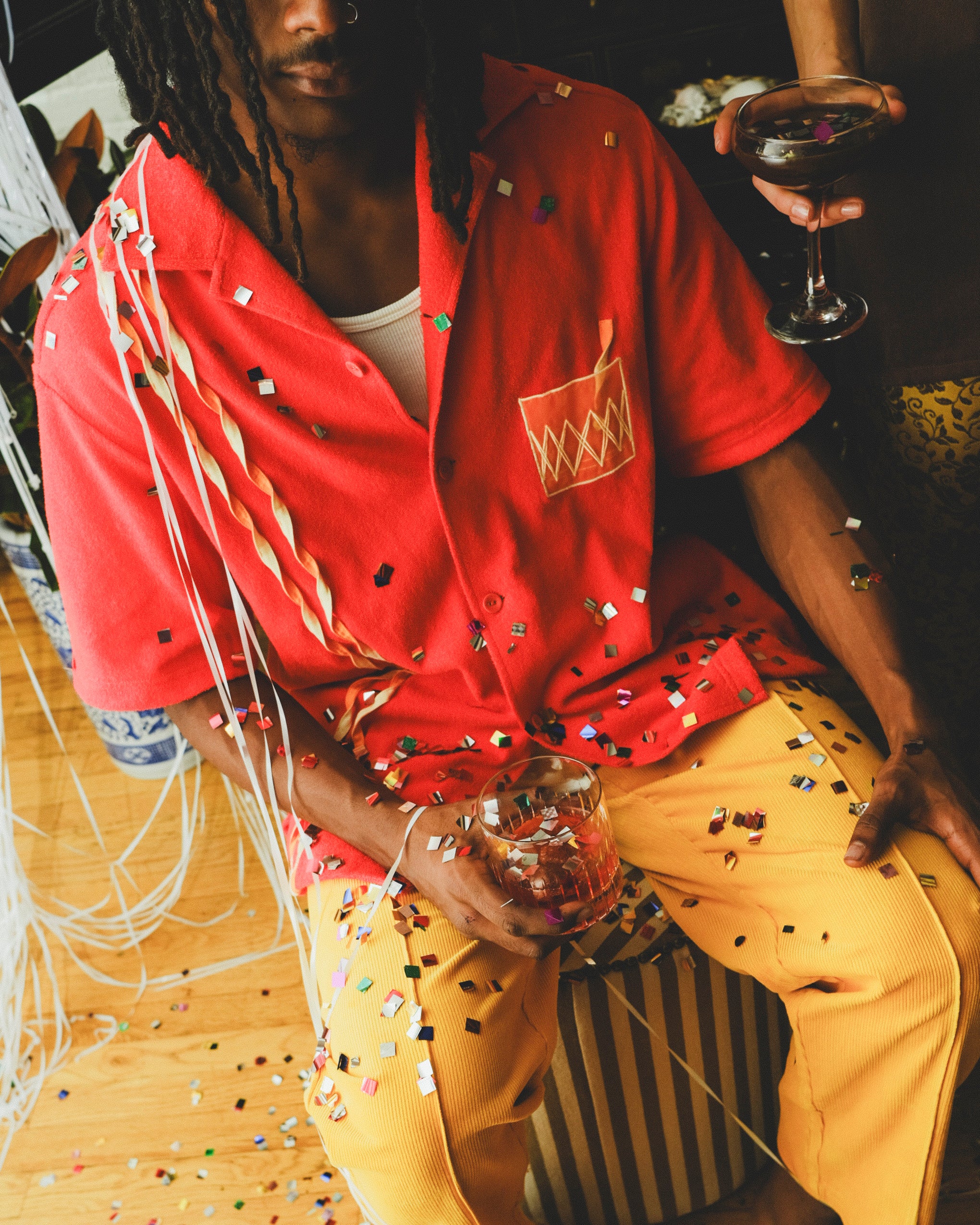 Model wearing a red cabana shirt with twisted appliqué stripes and an embroidered Negroni glass on the chest pocket, seated with confetti scattered across their clothing and surroundings. 
