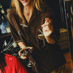 Smiling woman wearing a brown cabana shirt with espresso martini embroidery, holding a coupe glass filled with confetti. 