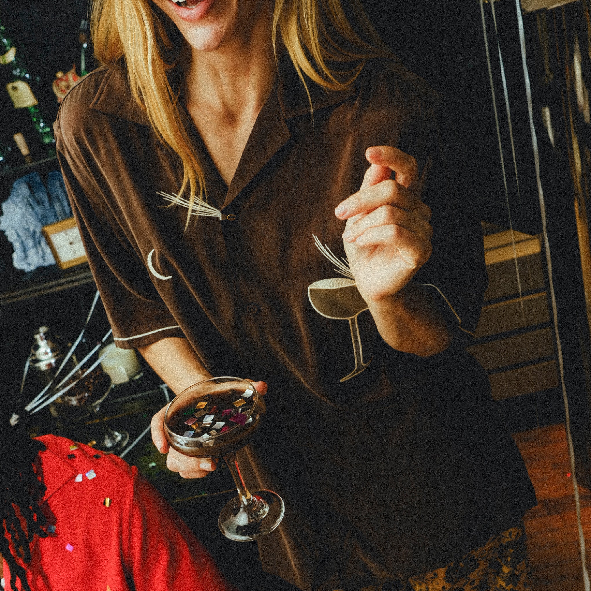 Smiling woman wearing a brown cabana shirt with espresso martini embroidery, holding a coupe glass filled with confetti. 