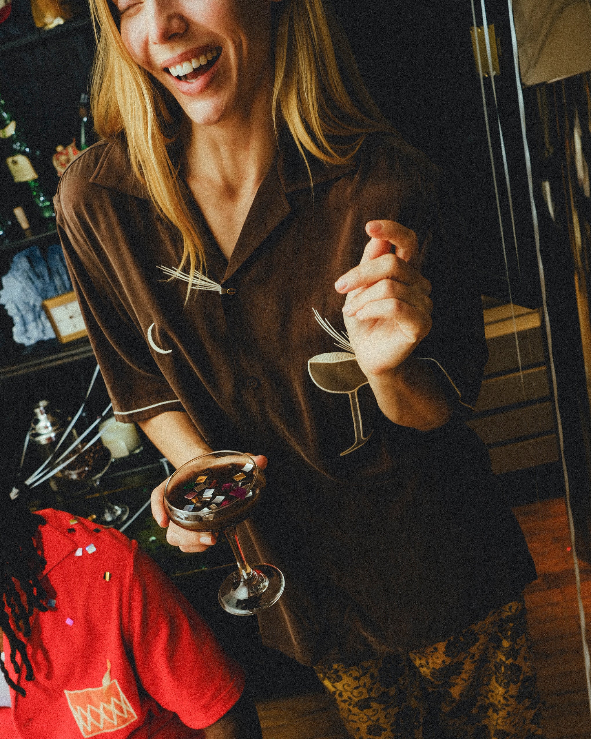 Smiling woman wearing a brown cabana shirt with espresso martini embroidery, holding a coupe glass filled with confetti. 