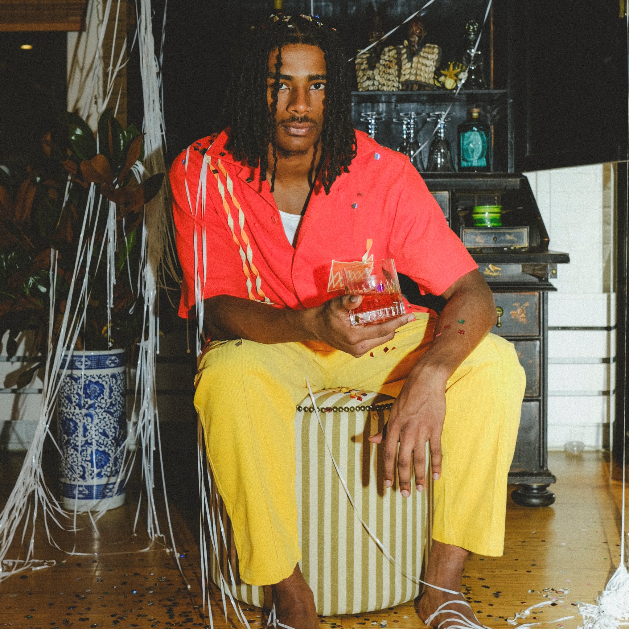Model wearing a red cabana shirt with twisted appliqué stripes and an embroidered Negroni glass on the chest pocket, seated on a striped ottoman. The model holds a Negroni-style drink in one hand.
