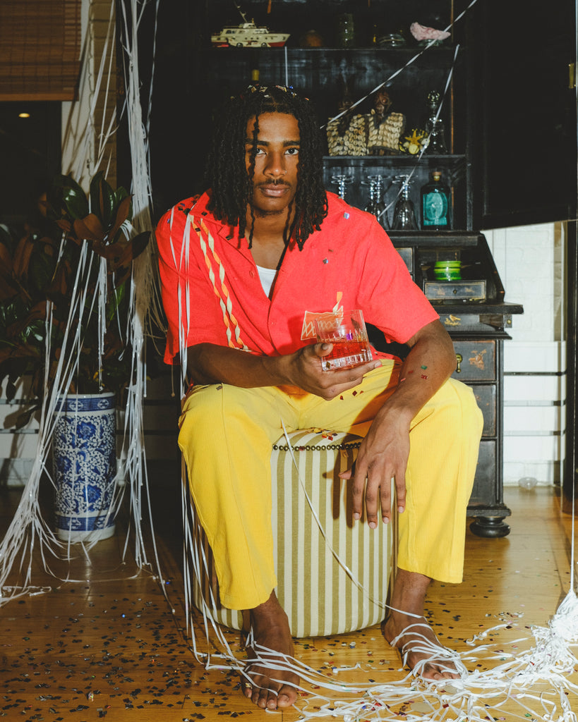 Model wearing a red cabana shirt with twisted appliqué stripes and an embroidered Negroni glass on the chest pocket, seated on a striped ottoman. The model holds a Negroni-style drink in one hand.