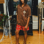 A man wearing a brown cabana shirt with cocktail-themed embroidery and cream piping, paired with red plaid shorts. 