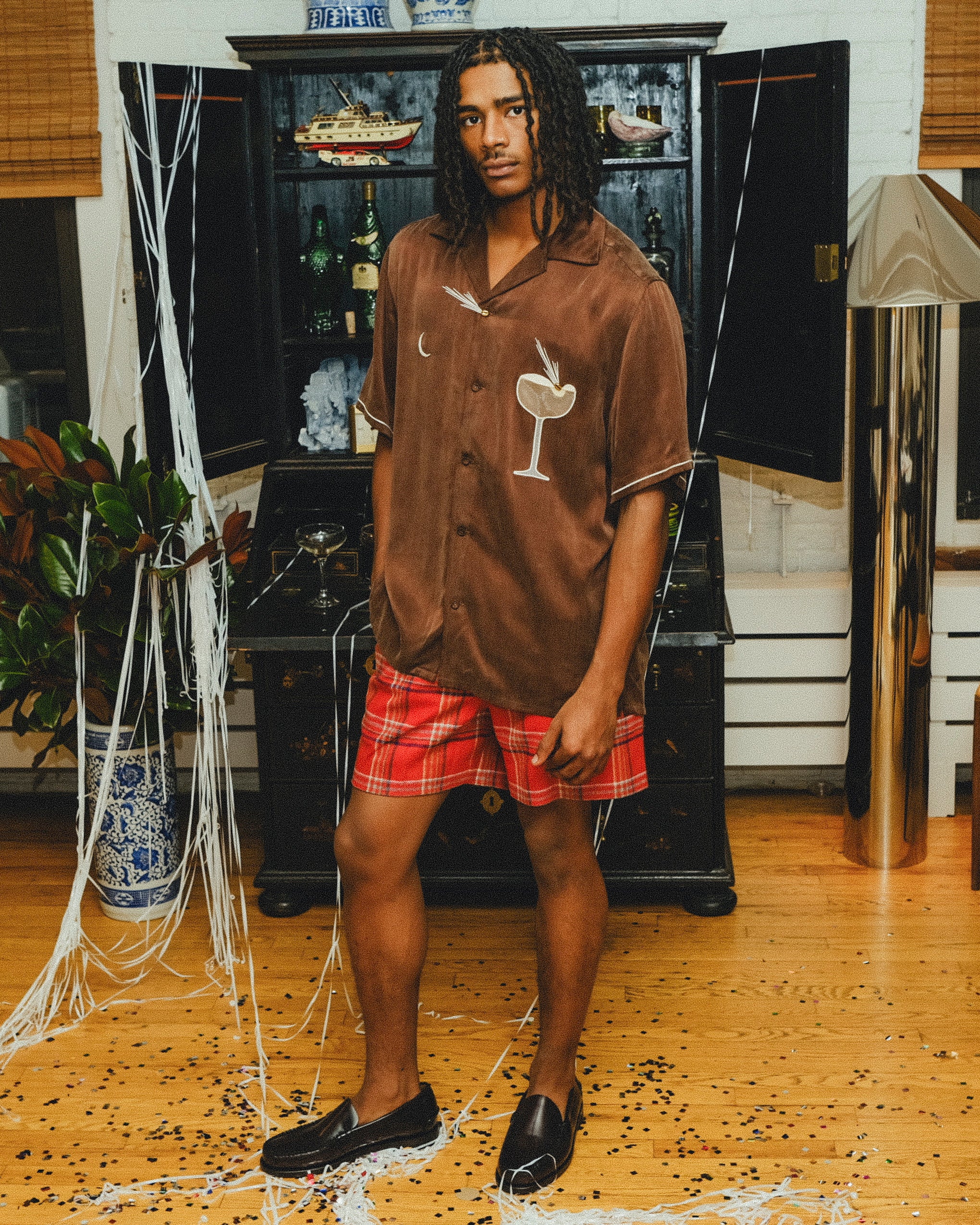 A man wearing a brown cabana shirt with cocktail-themed embroidery and cream piping, paired with red plaid shorts. 