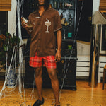 Man wearing a brown cabana shirt with cocktail-themed embroidery and cream piping, paired with red plaid shorts. 