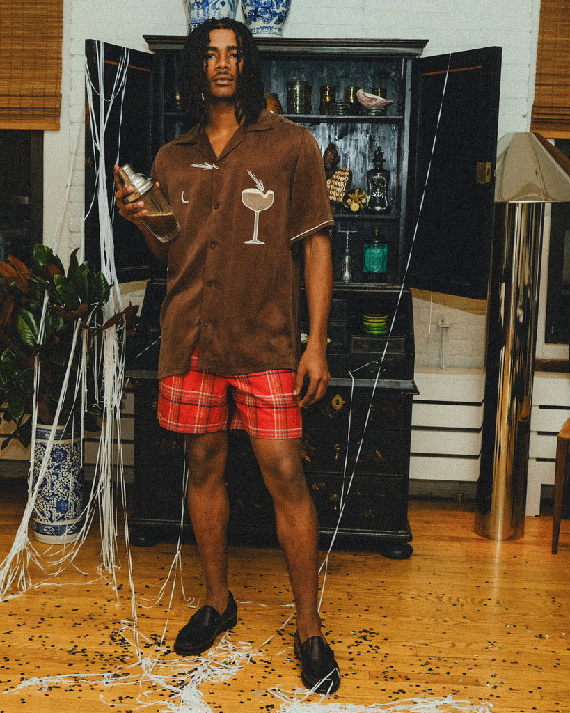 Man wearing a brown cabana shirt with cocktail-themed embroidery and cream piping, paired with red plaid shorts. 