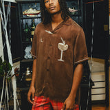 A man wearing a brown cabana shirt with cocktail-themed embroidery and cream piping, paired with red plaid shorts. 
