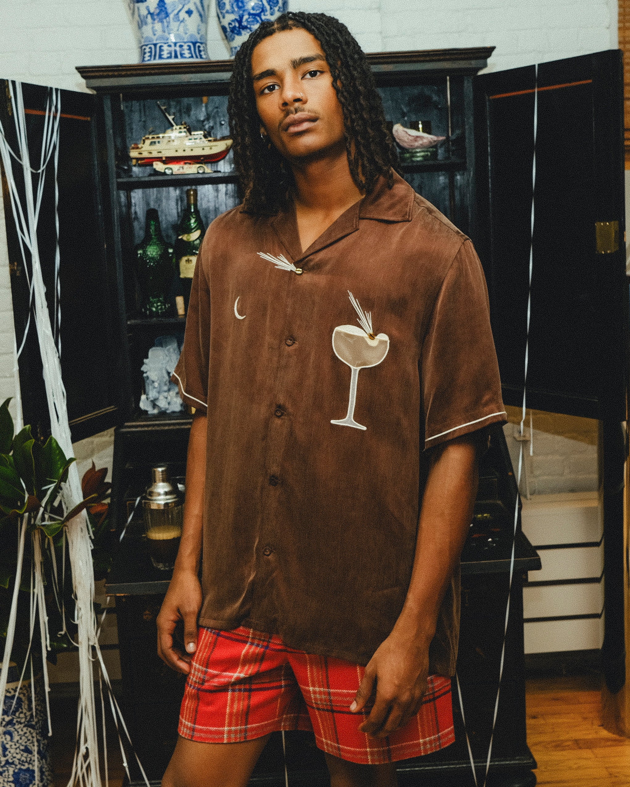 A man wearing a brown cabana shirt with cocktail-themed embroidery and cream piping, paired with red plaid shorts. 