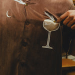 Close-up of a brown cabana shirt showing embroidered cocktail artwork, including a crescent moon and a coupe glass. A hand points to the garnish detail on the glass.