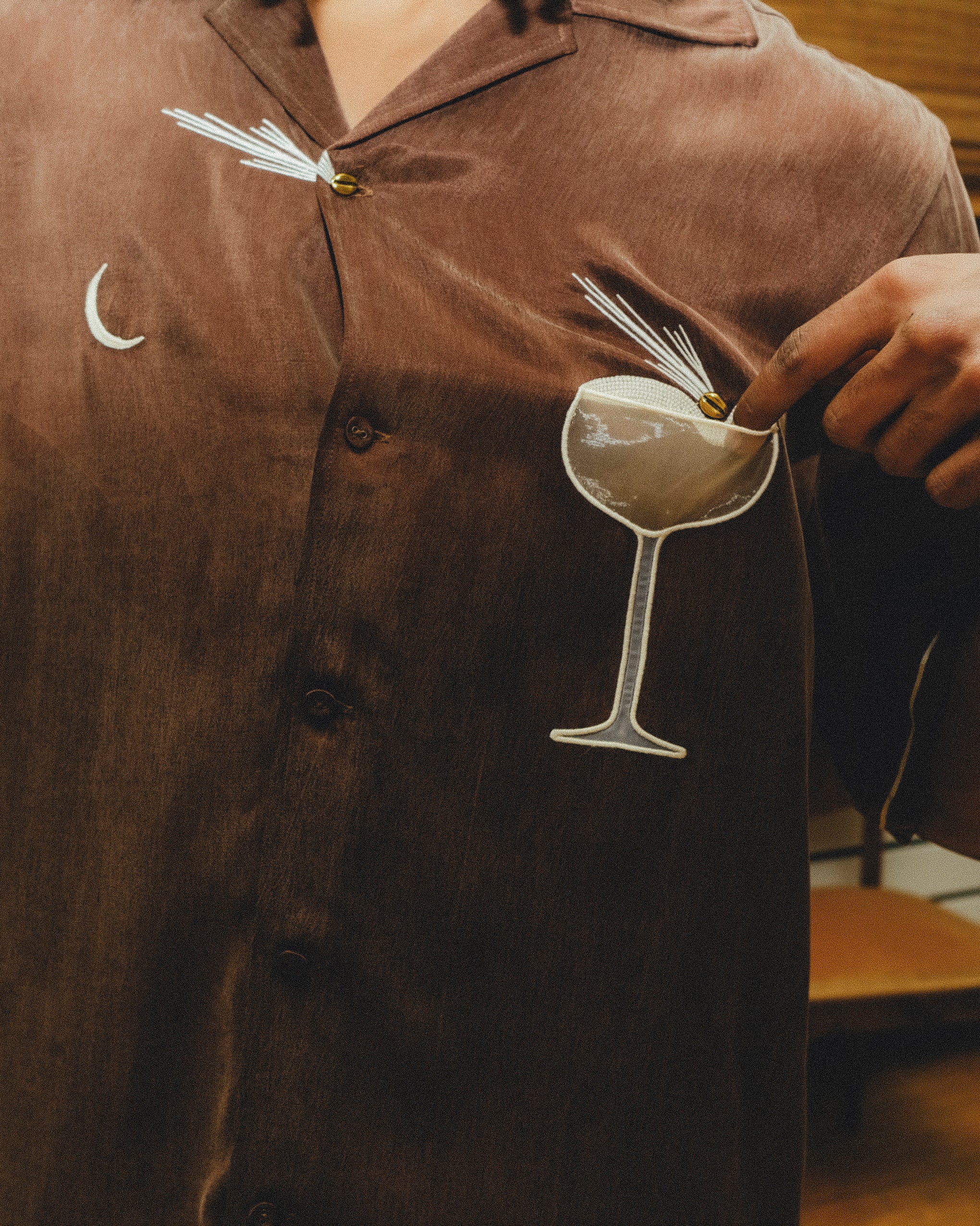 Close-up of a brown cabana shirt showing embroidered cocktail artwork, including a crescent moon and a coupe glass. A hand points to the garnish detail on the glass.