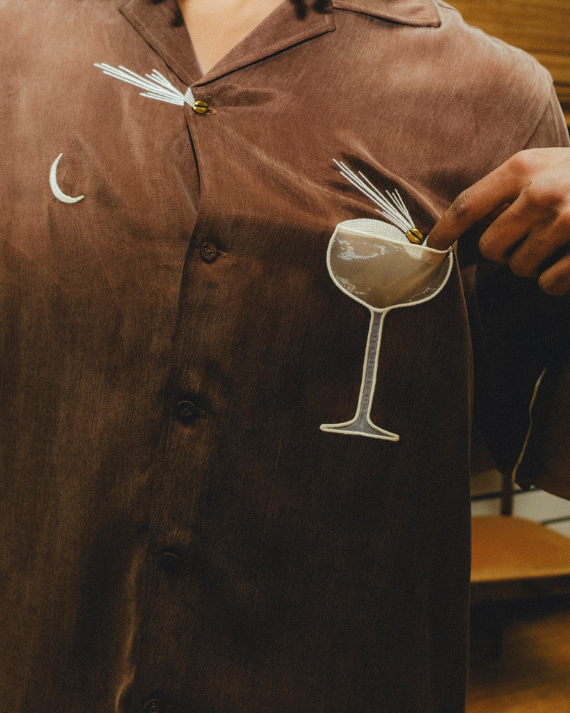 Close-up of a brown cabana shirt showing embroidered cocktail artwork, including a crescent moon and a coupe glass. A hand points to the garnish detail on the glass.