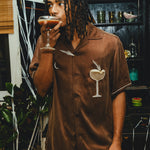 Man in a brown shirt with an espresso martini graphic, drinking from a glass in a bar setting.