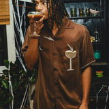 Man in a brown shirt with an espresso martini graphic, drinking from a glass in a bar setting.