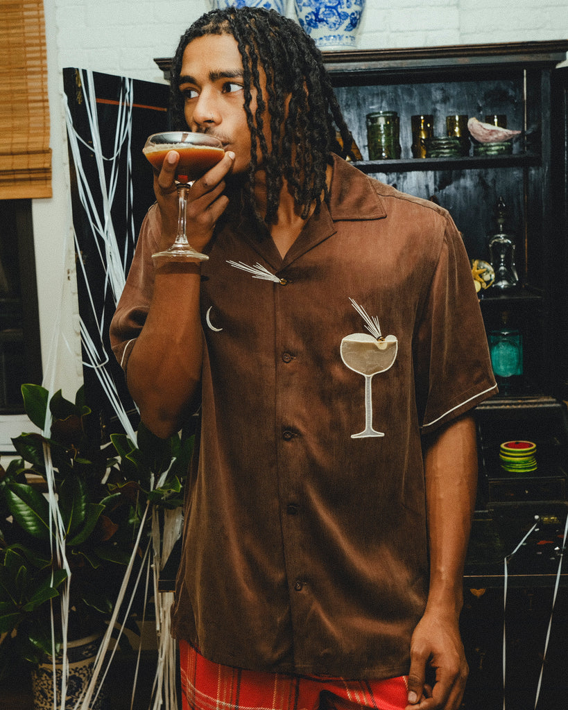 Man in a brown shirt with an espresso martini graphic, drinking from a glass in a bar setting.