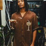 Man wearing a brown cabana shirt with cocktail-themed embroidery and cream piping, paired with red plaid shorts and holding an espresso martini in his hand. 