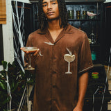 Man wearing a brown cabana shirt with cocktail-themed embroidery and cream piping, paired with red plaid shorts and holding an espresso martini in his hand. 