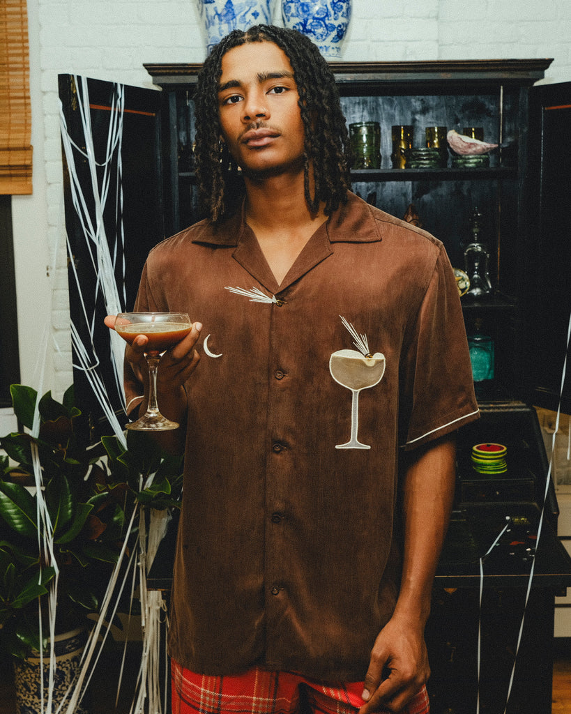 Man wearing a brown cabana shirt with cocktail-themed embroidery and cream piping, paired with red plaid shorts and holding an espresso martini in his hand. 