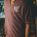 Model wearing brown polo shirt with 'Saturday Night Live' embroidery and yellow contrast embroidery.