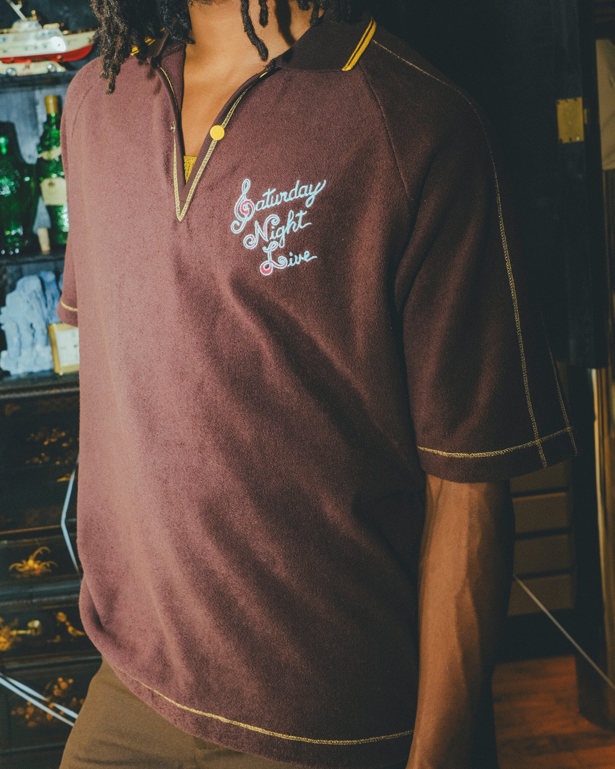 Model wearing brown polo shirt with 'Saturday Night Live' embroidery and yellow contrast embroidery.