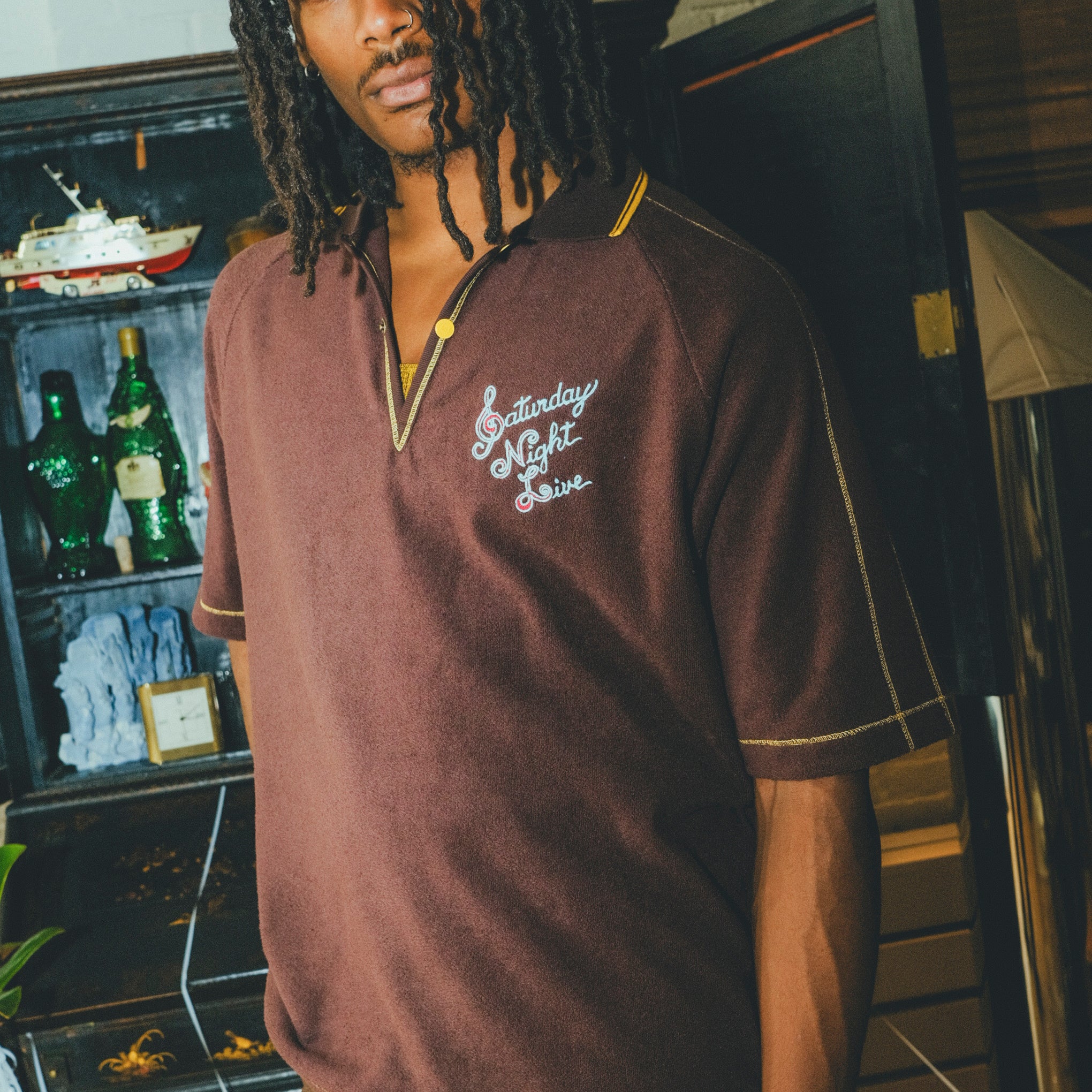 Man wearing a brown 'Saturday Night Live' polo shirt with yellow contrast embroidery.