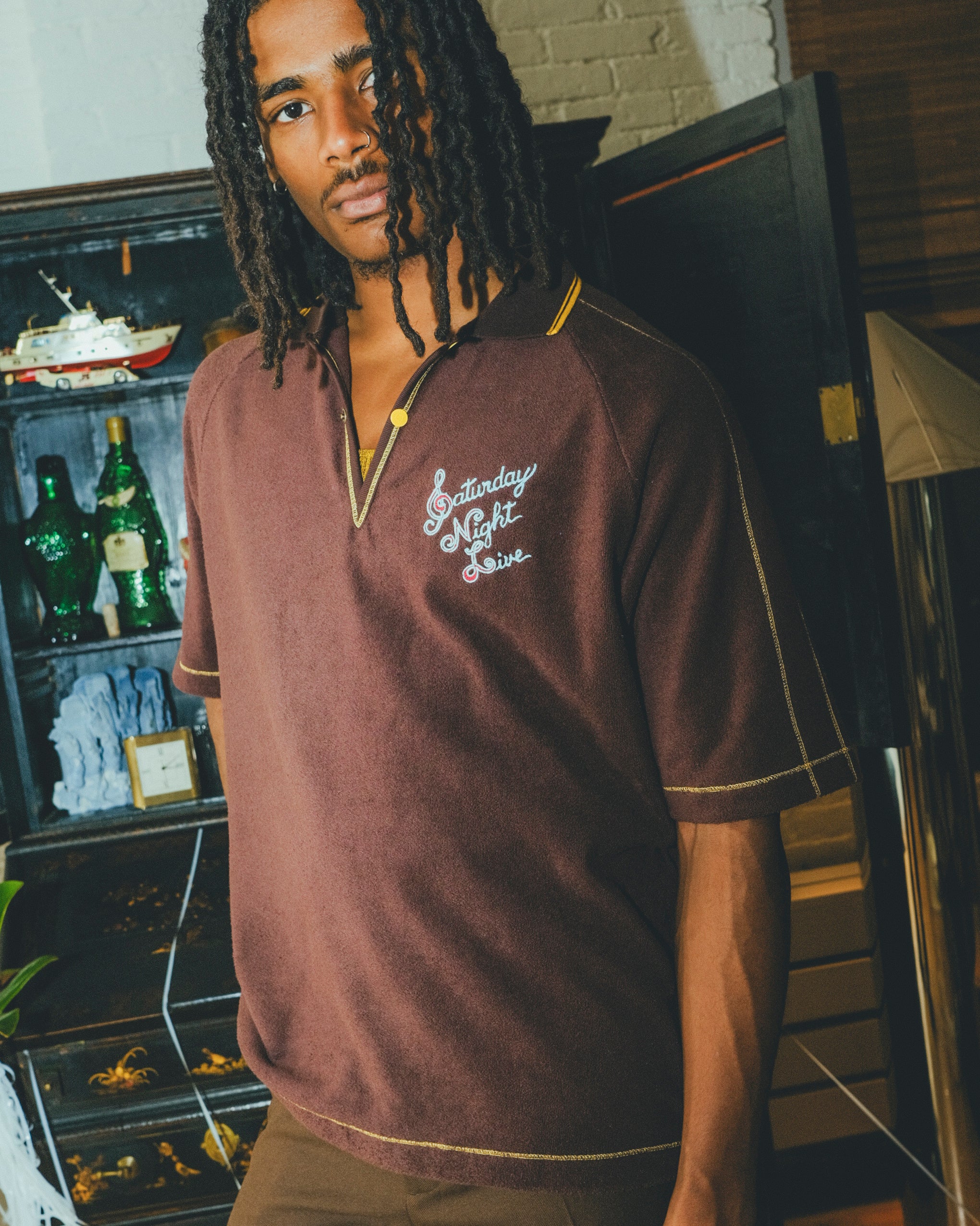 Man wearing a brown 'Saturday Night Live' polo shirt with yellow contrast embroidery.