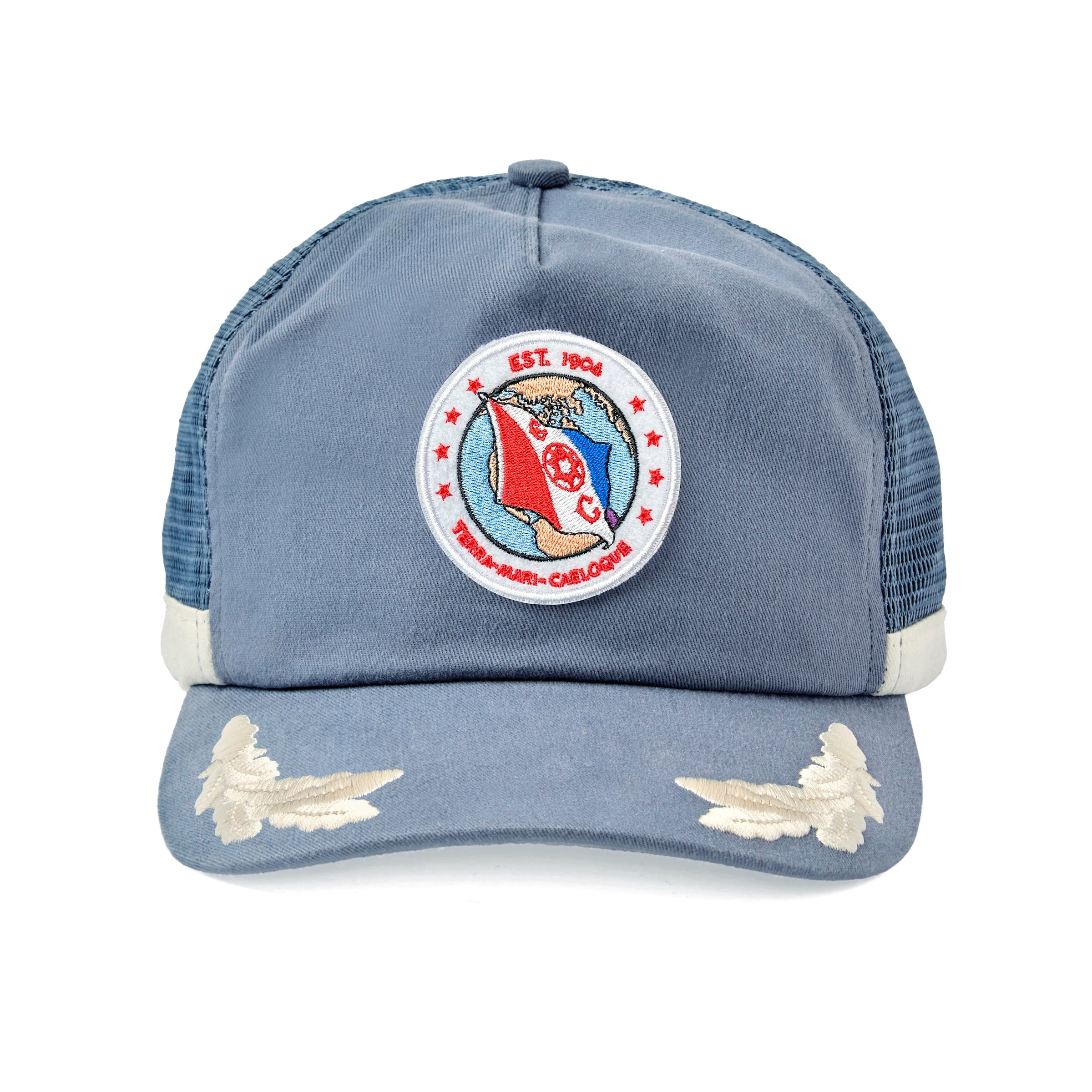 Blue hat with a circular embroidered patch on the front showing a red and blue flag over a globe. The patch reads “EST. 1904” at the top and “TERN-MARE-CAELOQUE” at the bottom, with the letters “E” and “C” on either side of a ship wheel. White embroidered wings decorate the brim.