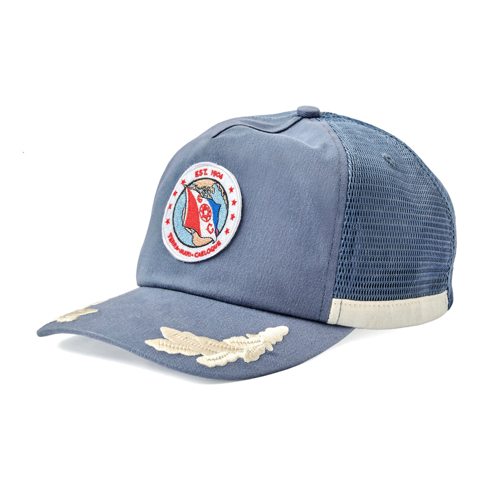 Blue mesh hat with a circular embroidered patch on the front showing a red and blue flag over a globe. The patch reads “EST. 1904” at the top and “TERN-MARE-CAELOQUE” at the bottom, with the letters “E” and “C” on either side of a ship wheel. White embroidered wings decorate the brim.