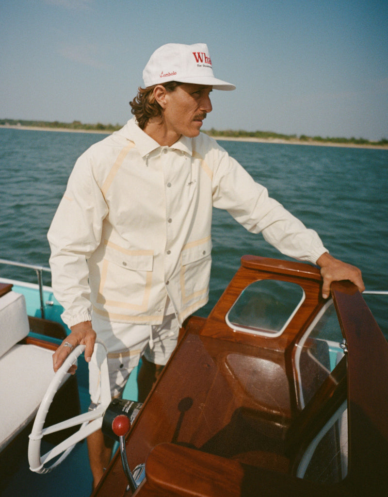 Man steering a boat while wearing a white five-panel cap with red text reading 'Whaler' and smaller text reading 'The Unsinkable Legend.' A small red 'Tombolo' logo is embroidered on the side of the hat. He is also wearing a light beige jacket and patterned shorts.