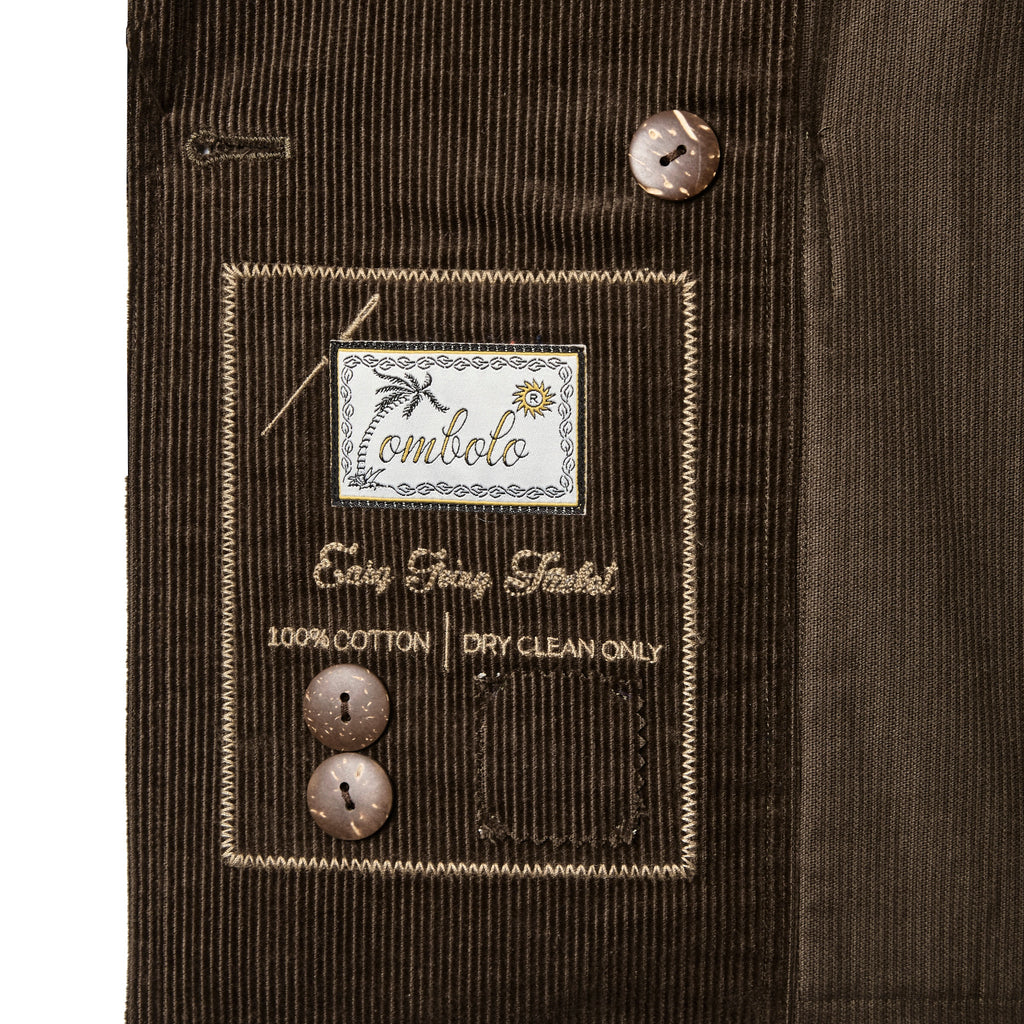 Close-up of the interior label on a brown corduroy jacket, featuring embroidered branding that reads 'Tombolo' and 'Easy Going Jacket' with care instructions and two spare brown buttons attached.