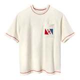 Cream-colored short-sleeve terry tee with red contrast stitching on the collar, sleeves, and hem. A chest pocket features a red and blue flag design with the letters “E” and “C” and a ship wheel emblem in the center.