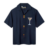 A navy blue short-sleeve button-down shirt with playful detailing: embroidered olive skewers act as buttons down the front, and a white martini glass with an olive garnish is embroidered on the chest. 