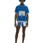 Model wearing a blue terry short-sleeve shirt with front graphic and mesh pocket detail, paired with a cream and blue FIFA World Cup bucket hat with chin cord.