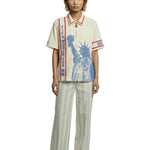 Model wearing a cream short-sleeve zip shirt with large blue Statue of Liberty graphic on the front, vertical FIFA World Cup lettering, and red contrast trim at the collar and sleeves.