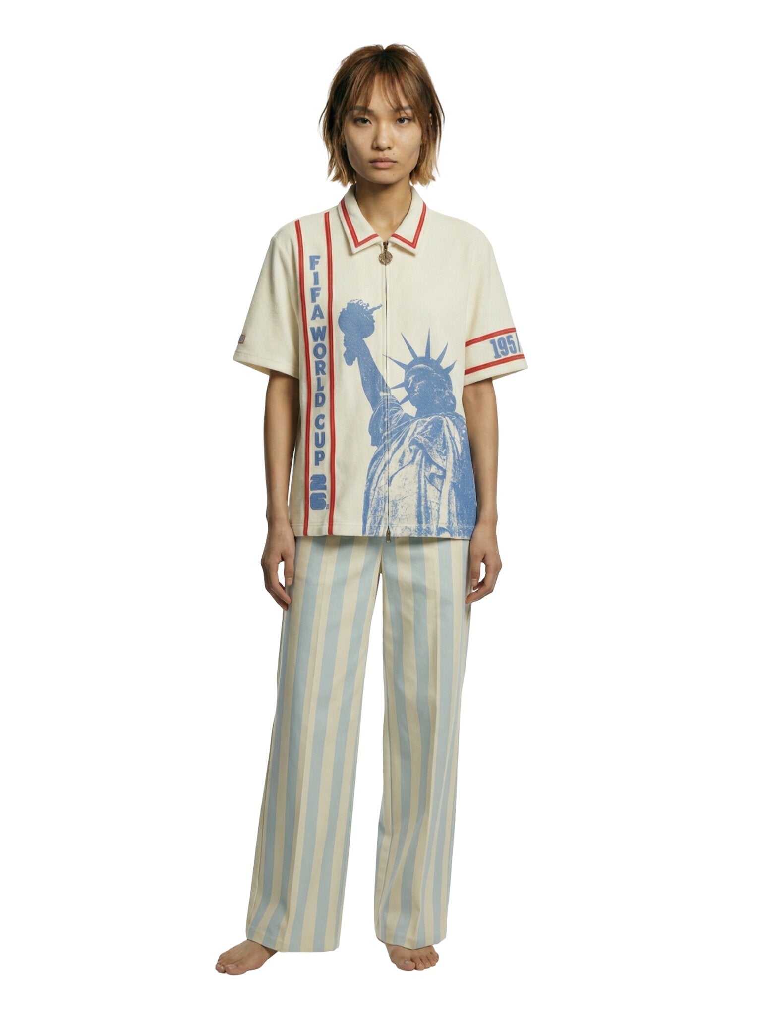 Model wearing a cream short-sleeve zip shirt with large blue Statue of Liberty graphic on the front, vertical FIFA World Cup lettering, and red contrast trim at the collar and sleeves.