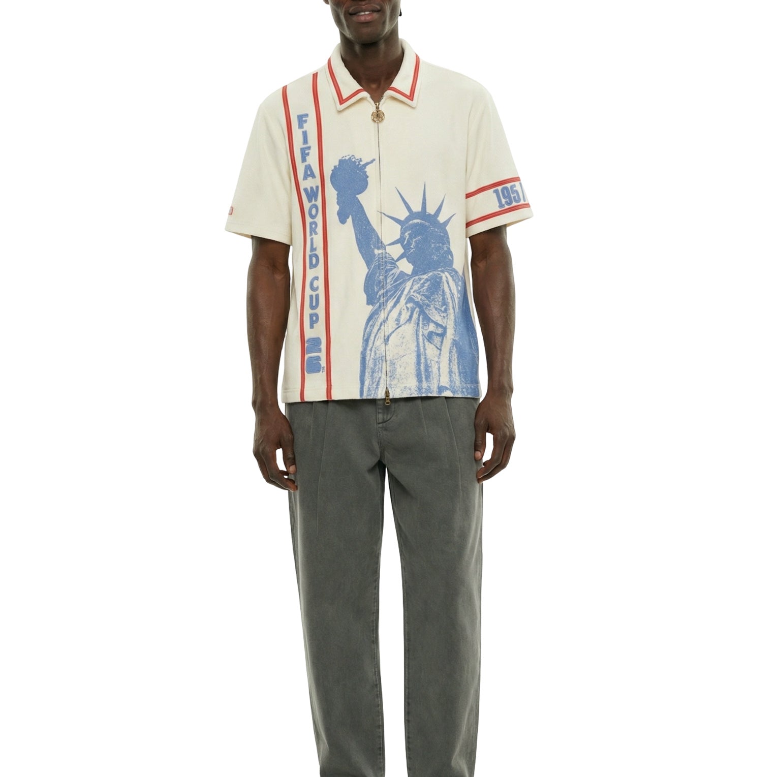 Model wearing a cream short-sleeve zip shirt with large blue Statue of Liberty graphic on the front, vertical FIFA World Cup lettering, and red contrast trim at the collar and sleeves.
