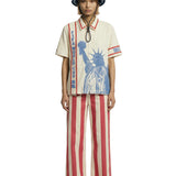 Model wearing a cream short-sleeve zip shirt with large blue Statue of Liberty graphic and vertical FIFA World Cup lettering, paired with a cream bucket hat featuring FIFA World Cup branding.