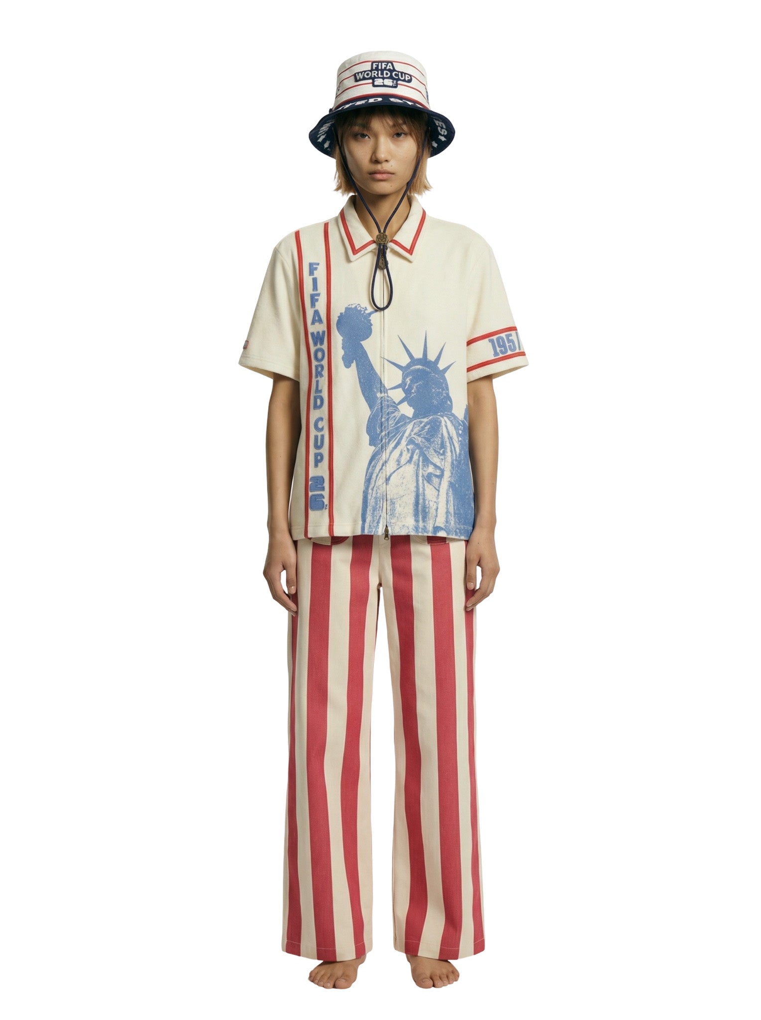 Model wearing a cream short-sleeve zip shirt with large blue Statue of Liberty graphic and vertical FIFA World Cup lettering, paired with a cream bucket hat featuring FIFA World Cup branding.