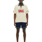 Man wearing a cream-colored shirt with 'USA' logo, navy shorts, and a matching hat on a white background