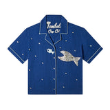 Front-facing product image of the 'Teamwork' Cabana ~ Crop Cut made of 60% Tencel and 40% Linen in a predominately navy coloring with a hand appliqué of a large fish chasing a smaller fish, fish-shaped agoya shell buttons serve as functional buttons on the placket and are scattered across the body, on the wearer's left panel, the fish-shaped buttons have been carefully assembled into the shape of a larger fish, chain-stitched interior label, and light-blue piping.