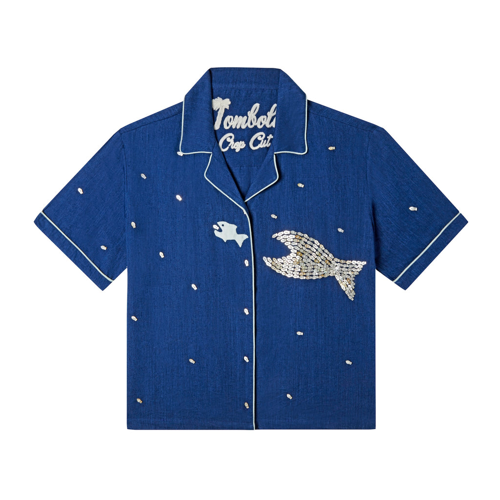 Front-facing product image of the 'Teamwork' Cabana ~ Crop Cut made of 60% Tencel and 40% Linen in a predominately navy coloring with a hand appliqué of a large fish chasing a smaller fish, fish-shaped agoya shell buttons serve as functional buttons on the placket and are scattered across the body, on the wearer's left panel, the fish-shaped buttons have been carefully assembled into the shape of a larger fish, chain-stitched interior label, and light-blue piping.