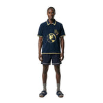 Model wearing a navy terry cloth FIFA World Cup short-sleeve shirt with yellow trim and a globe graphic on the front, paired with matching navy shorts.