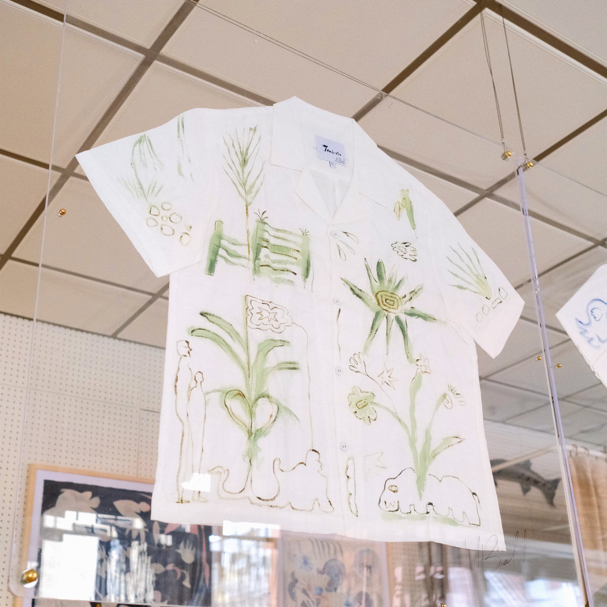 A short-sleeve white shirt with green botanical and abstract designs, displayed inside a glass case