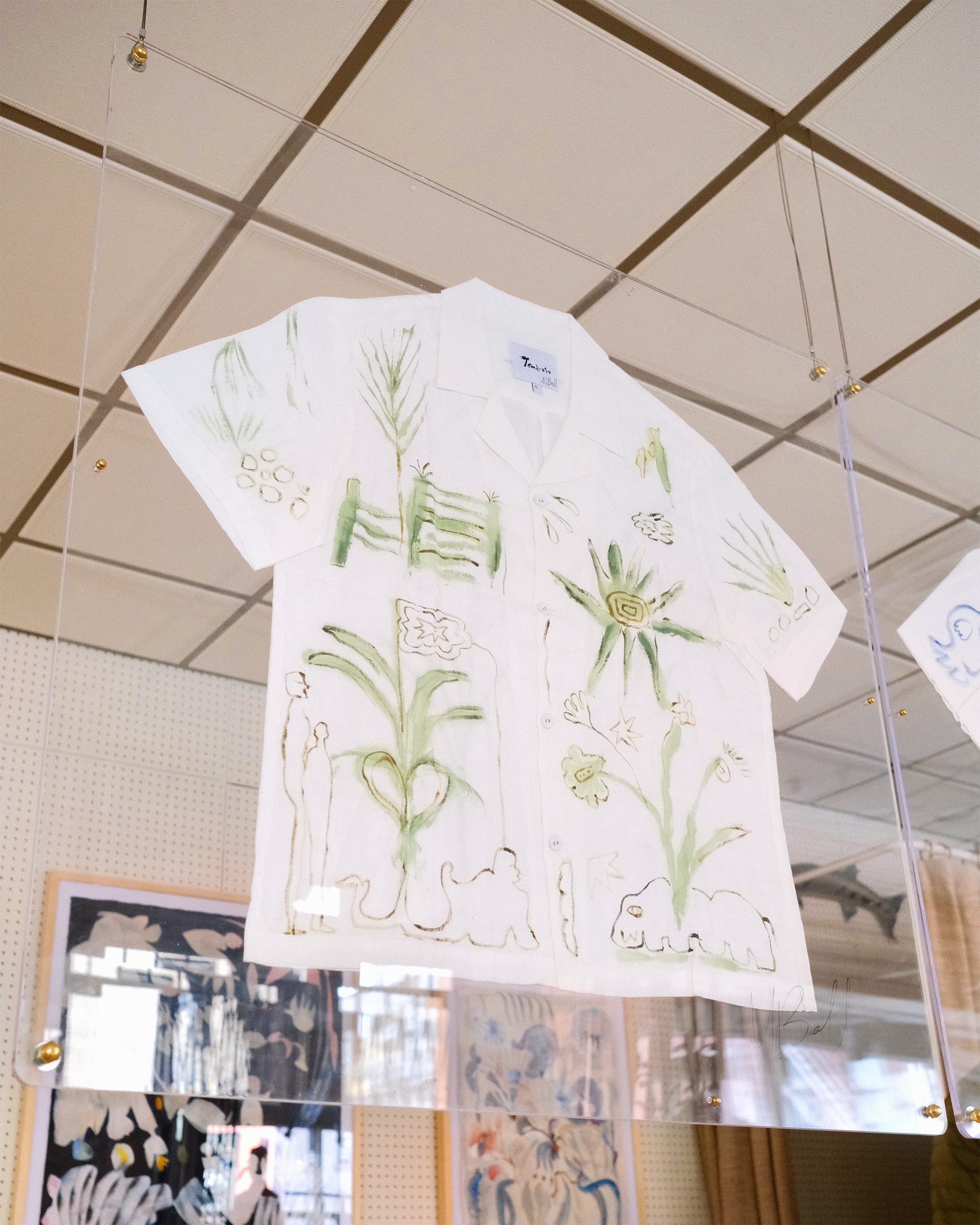 A short-sleeve white shirt with green botanical and abstract designs, displayed inside a glass case