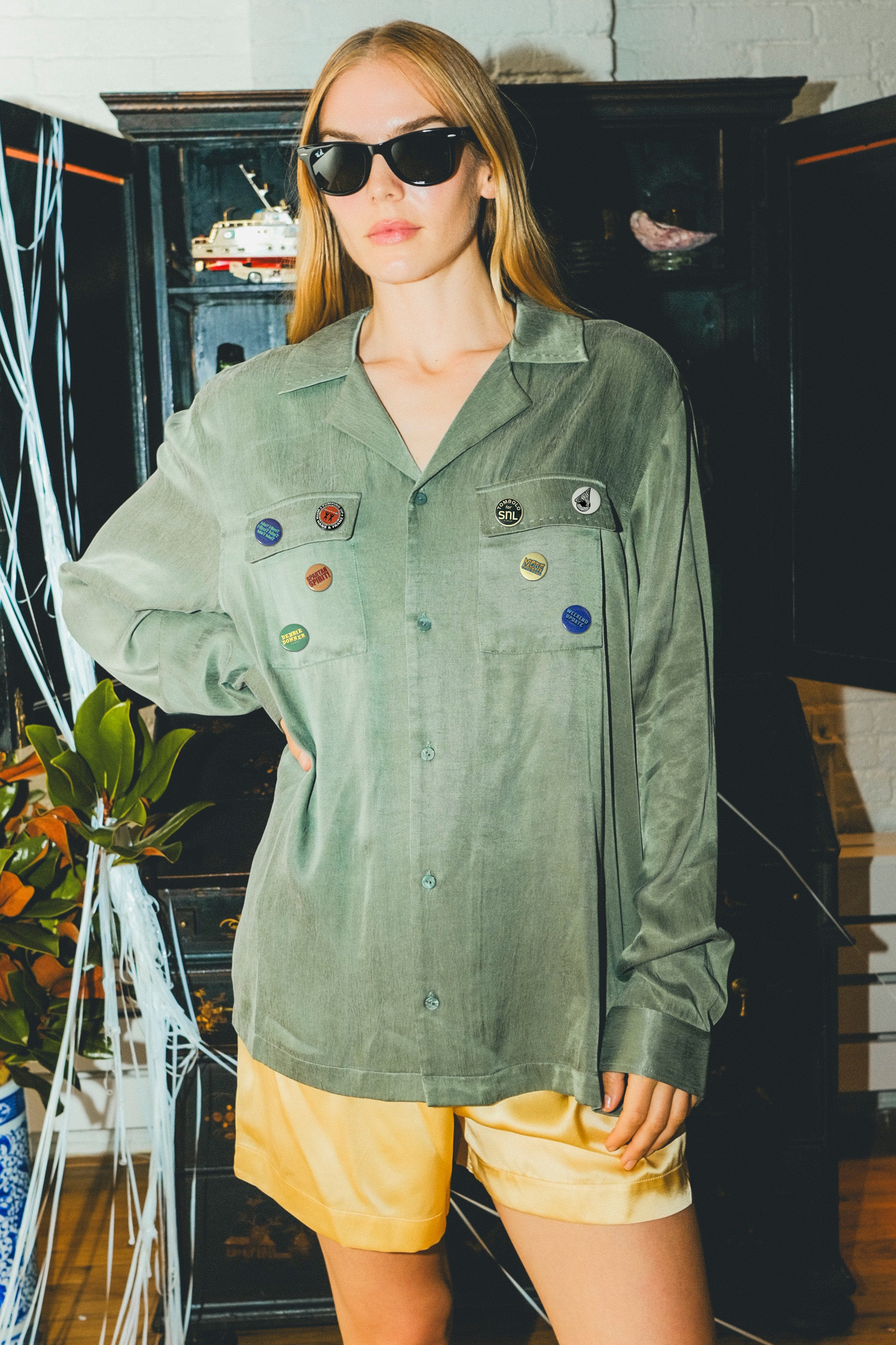 Woman wearing a green SNL shirt and sunglasses in front of a bar cabinet. 