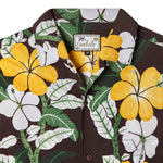 Close up front-facing product image of "Night Bloom" buttoned shirt with one button undone that emphasizes intricacies of hibiscus floral pattern that features yellow, green, and white accents on a brown base, in addition to the tag which shows a leaning palm tree as the "T" in Tombolo in a script font.